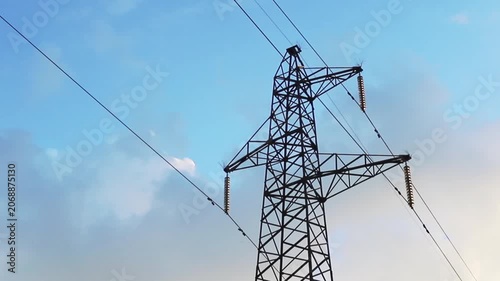 Metal electricity transmission tower stands against a blue sky with white clouds. High voltage power lines are suspended by insulators, carrying electricity across the landscape during daytime