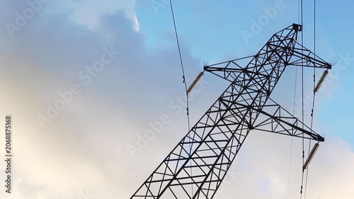 A sturdy industrial electricity transmission pylon rises against a clear blue sky, interspersed with soft white clouds