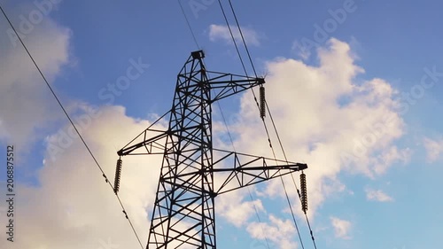 A large metal electricity pylon supports multiple high voltage power lines against a vibrant blue sky. Fluffy white clouds drift gently overhead on a bright day