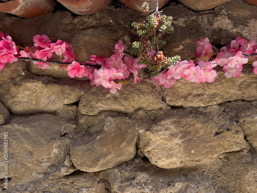 Horizontal close up eye level shot of a rustic ancient stone wall decorated with a garland of pink blossoms and a small hanging green succulent plant