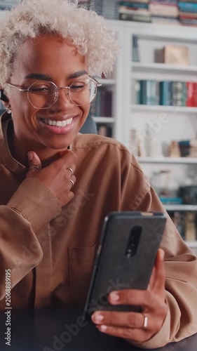 Cheerful African American woman with blonde curly hair and glasses holds her phone, chatting happily on a video call while wearing a wireless earbud in a bright indoor room.
