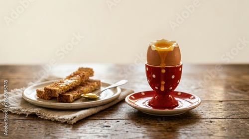 A perfectly soft-boiled egg with runny yolk dripping into a red polka dot egg cup alongside buttery toast on a wooden table
