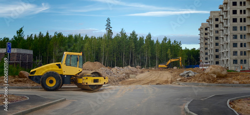 Yellow road roller and excavator on construction site with unfinished building and forest in the background under a clear blue sky