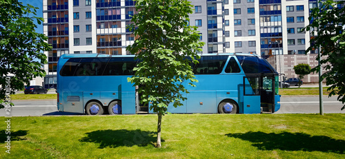 Blue tour bus parked beside a green lawn with trees in front of a modern residential building featuring large windows and a clear blue sky