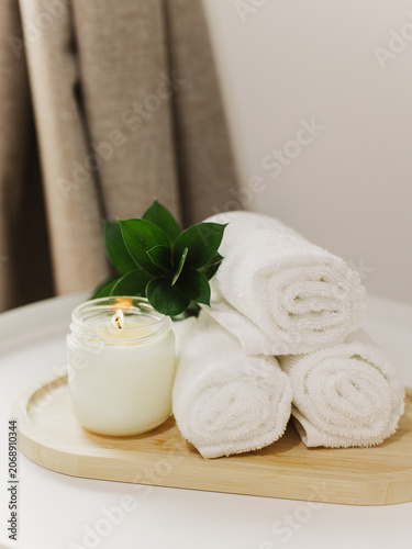 Minimalist still life of luxury spa essentials. White cotton towels with soy wax candle on a table.