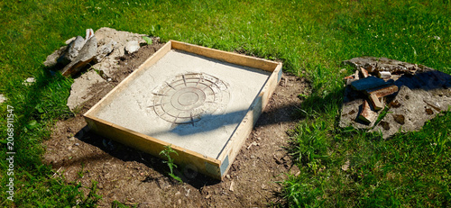 Concrete slab foundation with circular imprint surrounded by grass and debris, wooden frame visible, indicating preparation for construction or landscaping project