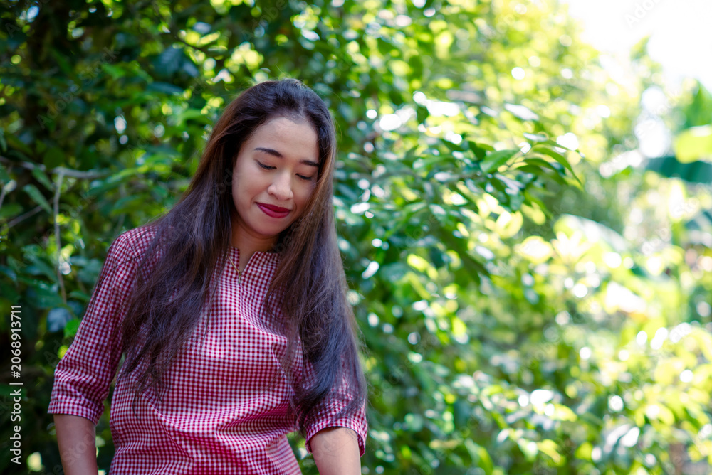 Fototapeta premium A young woman smiles softly beneath the green leaves. Her pink plaid shirt adds a cheerful contrast.