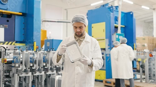 Young worker wearing protective uniform and hairnet smiling while holding stack of aluminum containers. Standing in modern industrial factory. Clean production environment indoors.