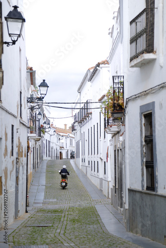 A narrow cobblestone street lined with traditional whitewashed houses and vintage iron lanterns as a person on a scooter drives away in the historic old town of Zafra, Badajoz, Spain.