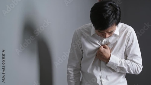 Experiencing chest pain, Asian man doubling over holding chest by gray wall, white shirt and shadow