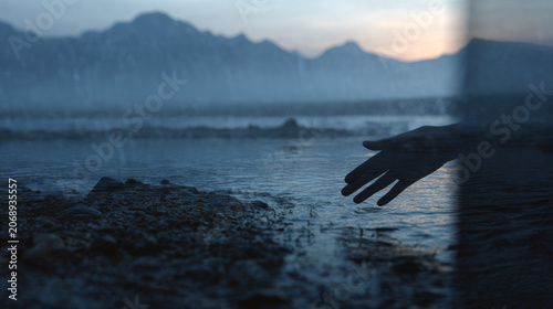 Silhouette of a hand reaching toward calm mountain lake water during twilight with a subtle glass reflection effect.
