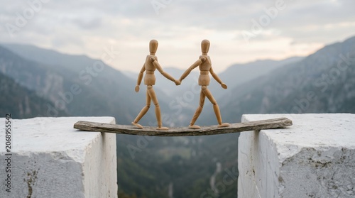 Two wooden figures holding hands on narrow plank between stone blocks, symbolizing trust, balance, and connection against scenic mountain valley under cloudy sky.