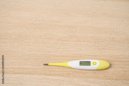 Green yellow digital thermometer lying on a light wooden table, used for measuring body temperature, fever monitoring and home health care. Simple health, illness and personal care concepts.
