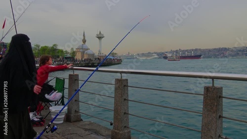 A woman wearing a black niqab holds a fishing rod while sitting on the stone embankment of Uskudar overlooking the Bosphorus sea and mosque in Istanbul