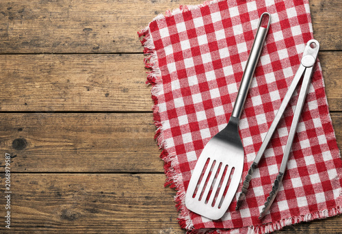 Stainless steel spatula and tongs on red checkered cloth on wooden table