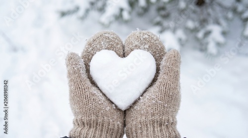Hands in beige knitted mittens holding a heart shaped snowball in a snowy forest