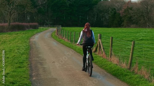 Middle aged woman cycling countryside road between green meadows and forest edge enjoying outdoor fitness. Active female cyclist riding bicycle along rural lane during calm daylight promoting healthy