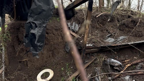 Collapsed remains of a destroyed military dugout in a leafless forest — charred beams, caved-in earth walls, torn sheeting and debris in mud. Aftermath of war; early spring, overcast