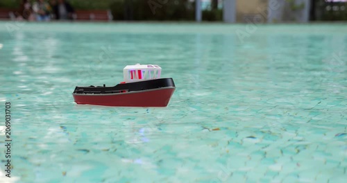 Miniature red tugboat with white cabin sways gently in place on calm turquoise water of model boat pool at Victoria Park, Hong Kong. Serene close-up captures charm of this beloved local hobby.