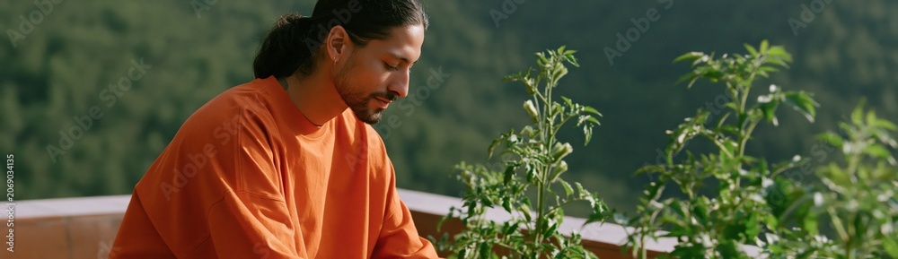 Naklejka premium Man in orange sweater gardening on balcony with green plants overlooking forested hills for urban nature and wellness background