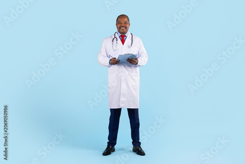 Foto Confident mature African American doctor in white coat with stethoscope, holding