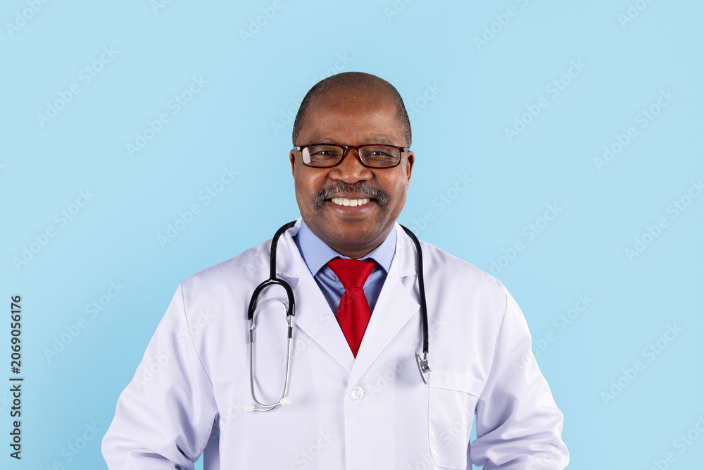 Fototapeta premium Closeup portrait of smiling mature African American doctor wearing glasses, white coat, stethoscope and red tie, blue studio background with copy space