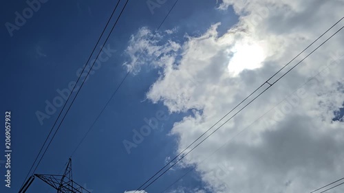 A timelapse captures white clouds moving across a blue sky. Overhead pylon wires stretch over the horizon, bridging nature and technology. The shifting climate transforms a bright day into an overcast