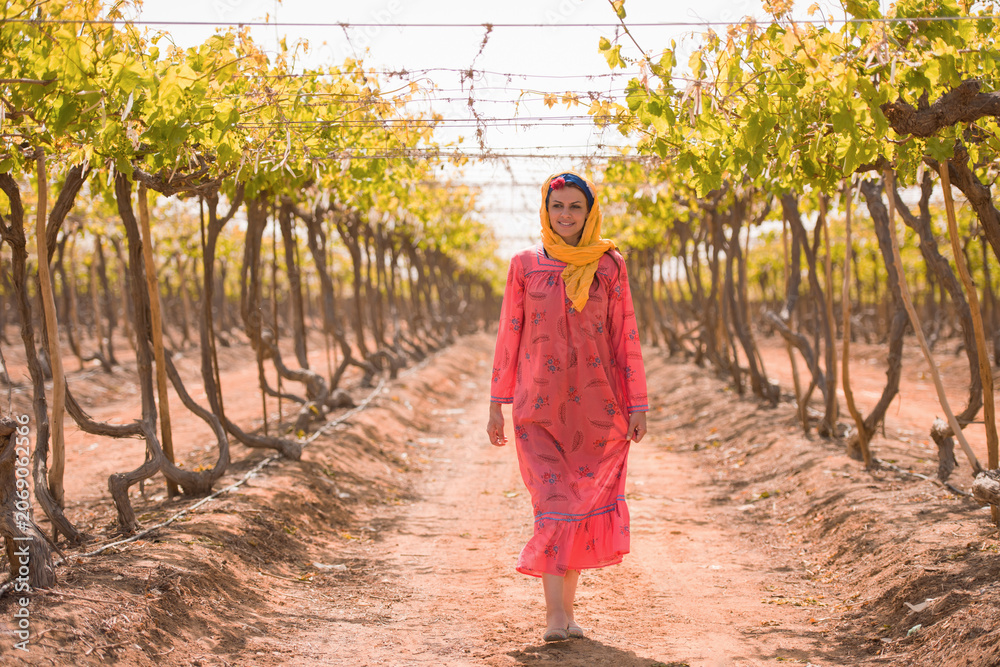 Fototapeta premium Portrait of confident Middle Eastern woman agricultural worker in traditional embroidered pink dress and yellow headscarf standing in symmetrical rows
