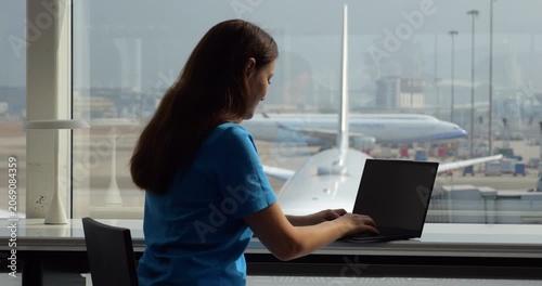 Passenger woman opens her laptop and begins working while seated at table by airport terminal windows. Focused and purposeful, she types on keyboard, making productive use of time before her flight.