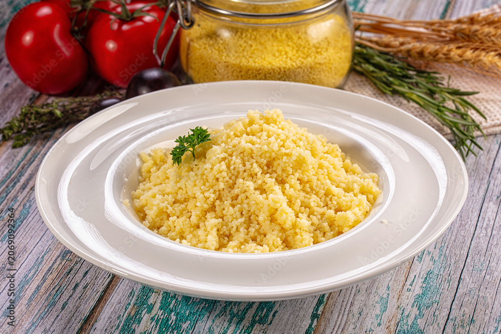 Fototapeta premium Cooked couscous on white plate with fresh tomatoes and herbs on rustic wooden table