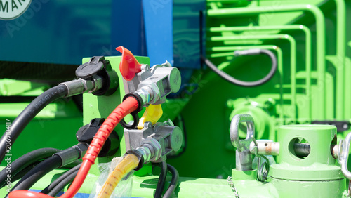 Close-up view of bright green industrial machinery showing metal connectors, thick hoses, colored cables, and mechanical fittings with strong daylight and blurred equipment in