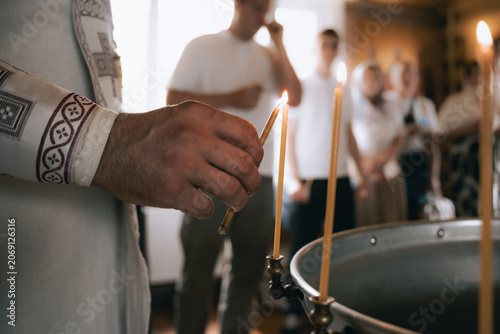 Quadro su tela Clergy hand lighting slender candles beside a metal baptismal font during a solemn church ceremony, with blurred congregants observing the sacred ritual in a reverent interior
