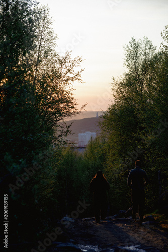 Two people walking down wooded path toward distant city at sunset