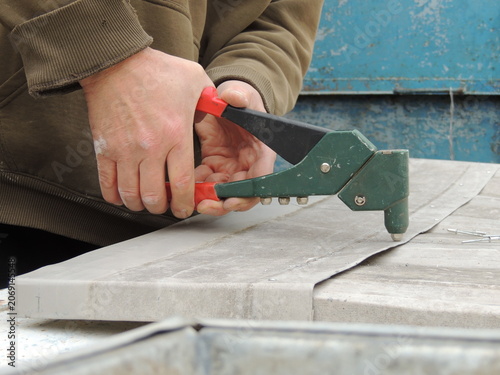 The technician holds a hand-operated lever riveter and squeezes the tool's handles to secure the fastener. The connection is made on curved sheets of thin gray metal.