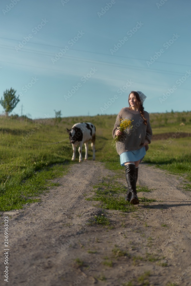 Fototapeta premium Woman walking along a dirt path with a cow in a lush green field under a blue sky during a sunny day