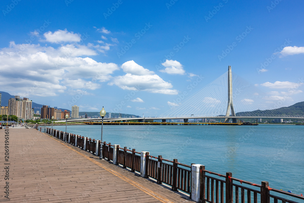Fototapeta premium Wooden seaside walkway overlooking modern Danjiang Bridge in Tamsui Taiwan
