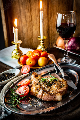 Tableau sur toile Romantic candlelit dinner featuring a perfectly grilled pork neck steak on a vintage metal platter, served with red wine, fresh rosemary, and ripe tomatoes in a warm, rustic atmosphere