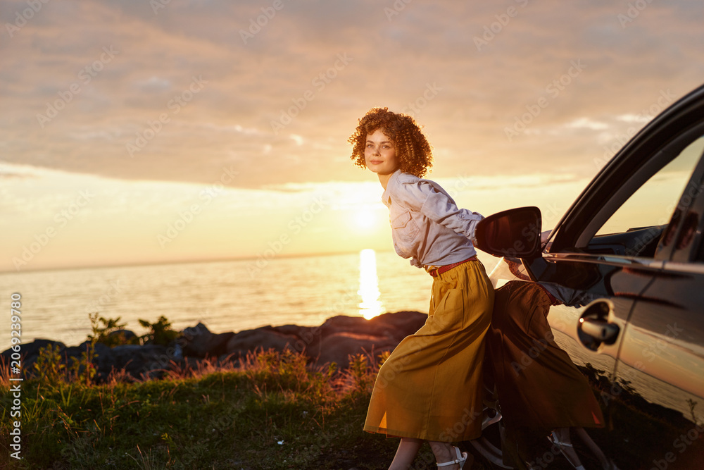 Fototapeta premium A young woman with curly hair leans on a dark car at sunset by a rocky coastline, conveying a moment of travel and quiet confidence.