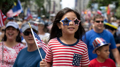 Happy young girl in patriotic clothing holding an American flag at a street parade, celebrating Independence Day or Memorial Day with family and community during summer