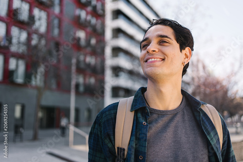 Portrait young hispanic man with backpack standing in the city looking to the side with confident expression