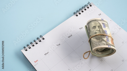 Roll of US dollar bills sitting on top of a monthly desk calendar planner concept of financial deadlines tax day payment schedule or savings