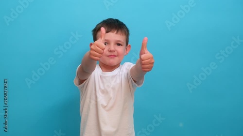 Little boy showing thumb up gesture on blue background. Happy child expressing positive emotion, approval and success. Studio portrait of youngster wearing white t-shirt, gesture concept.