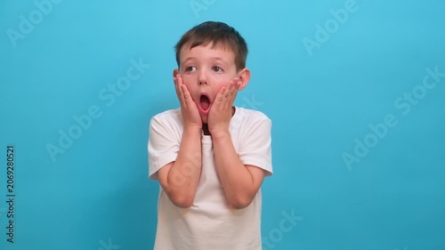 Surprised boy holding face with hand. Shocked kid with mouth open looking at camera. Emotional child expressing disbelief and amazement on solid blue background. Human facial expression.