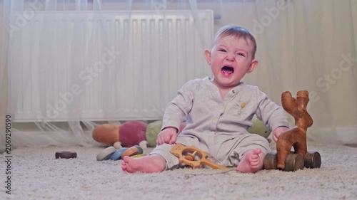 Crying baby boy sitting on light floor with wooden toy and rattle. Sad infant feeling upset at home. Emotional childhood expression, development and nursery interior concept.