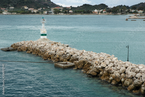 A small white and green lighthouse stands at the end of a long rocky stone breakwater protecting the harbor of Zakynthos Town in Greece
