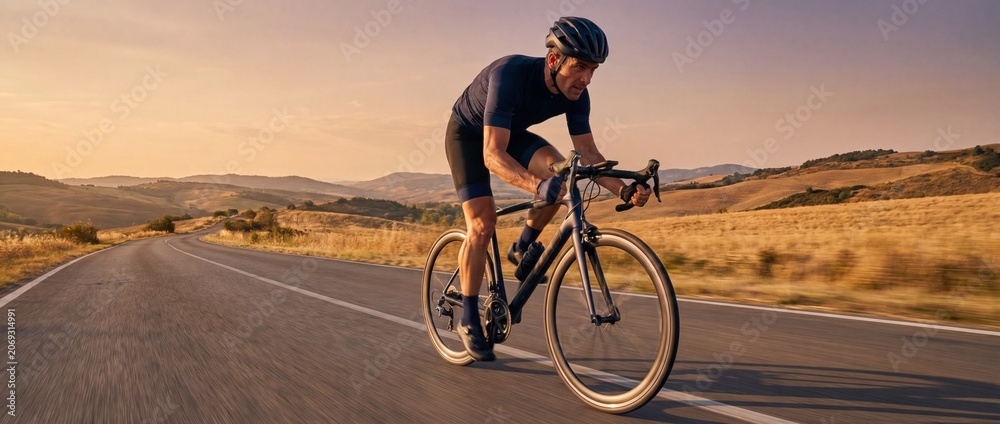Fototapeta premium Athletic male cyclist riding road bike on scenic winding asphalt road through rolling hills during golden hour sunset capturing speed