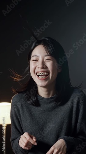 Cheerful young Asian woman with dental braces laughing sincerely while looking at camera on dark atmospheric studio background, genuine happiness concept, portrait video style.