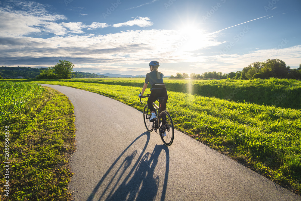 Fototapeta premium Female cyclist with road bike riding on asphalt path at sunset in summer. Slovenia. Rear Woman cycling in helmet on bicycle lane near green fields, active sport lifestyle, sun sky landscape. Lifestyle