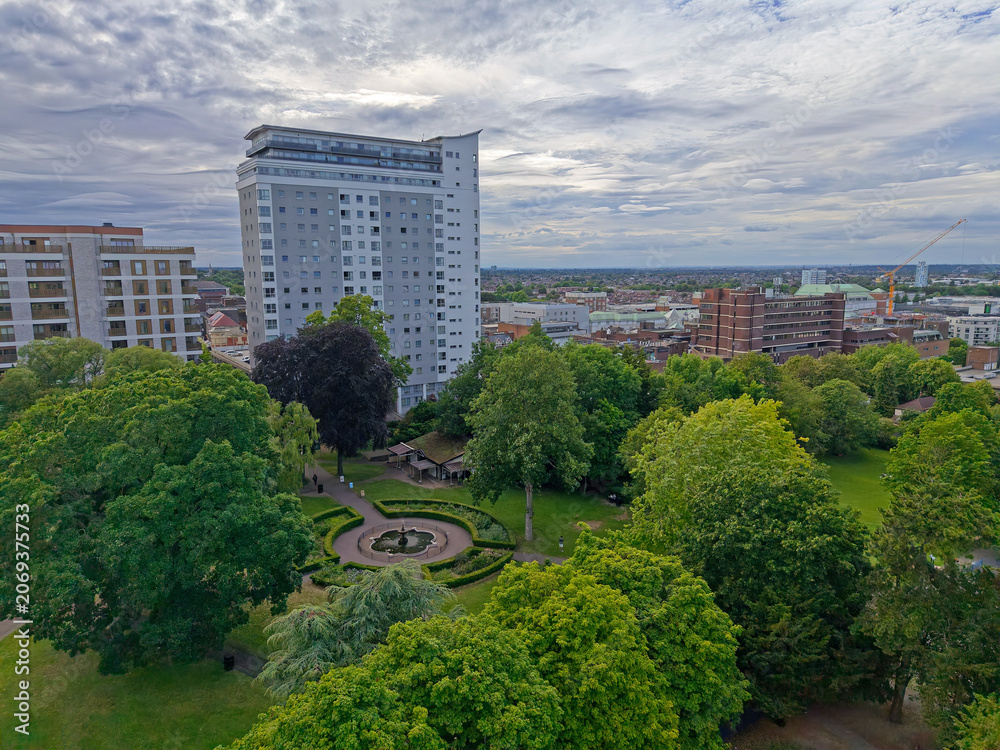 Fototapeta premium Manor park in Sutton town center, Greater London, UK. Aerial view above green public park with trees on a cloudy day. High rise apartment building. View towards high street with shopping center.