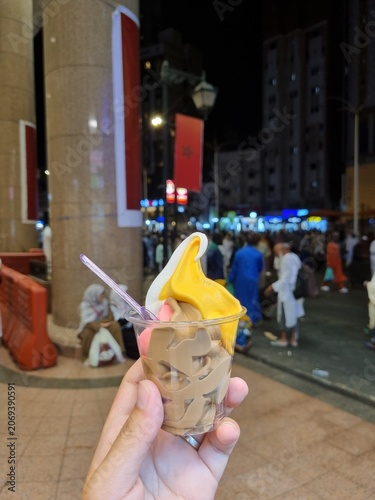 Bild auf Leinwand A hand holding a plastic cup filled with colorful soft-serve ice cream, set against a blurry, bustling night street scene